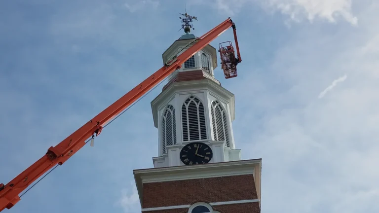 Worker in aerial lift basket inspecting white church steeple with clock and weathervane