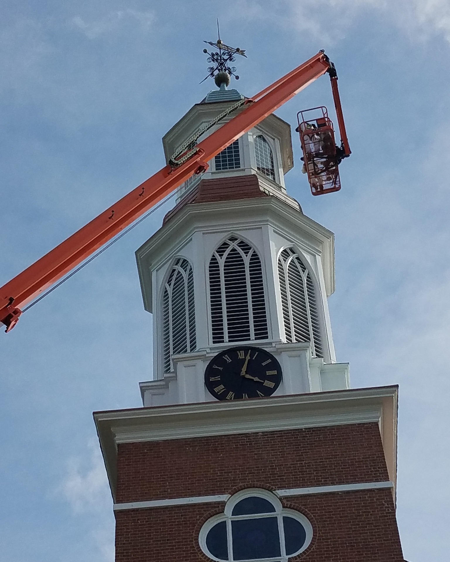 Orange boom lift extended to white church steeple with clock tower and weathervane