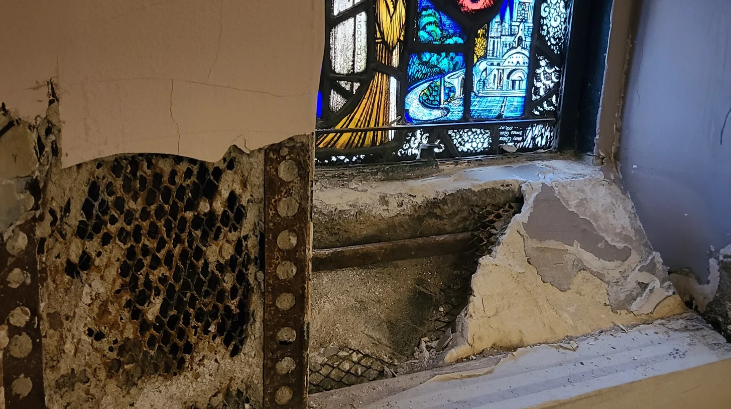 Deteriorated wall plaster and water damage below a stained glass window in a church