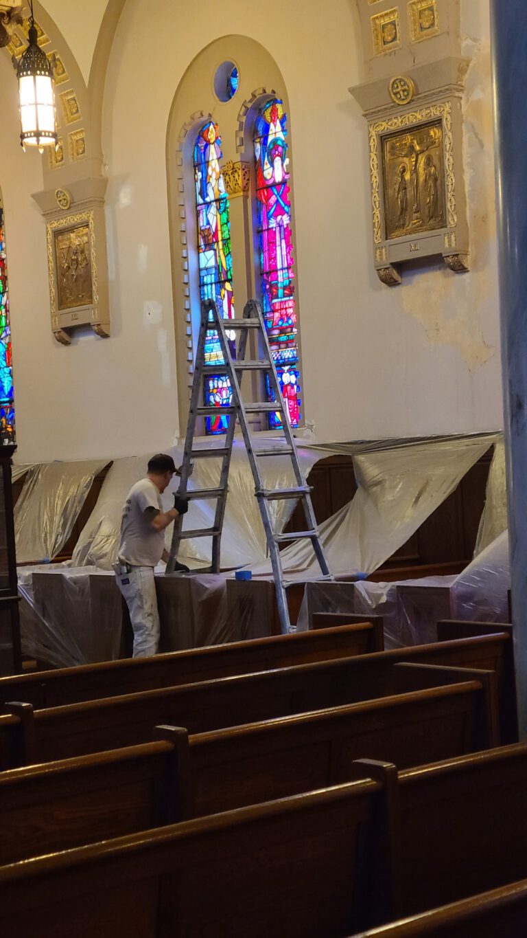 Worker on ladder beside draped pews in church nave near stained glass windows