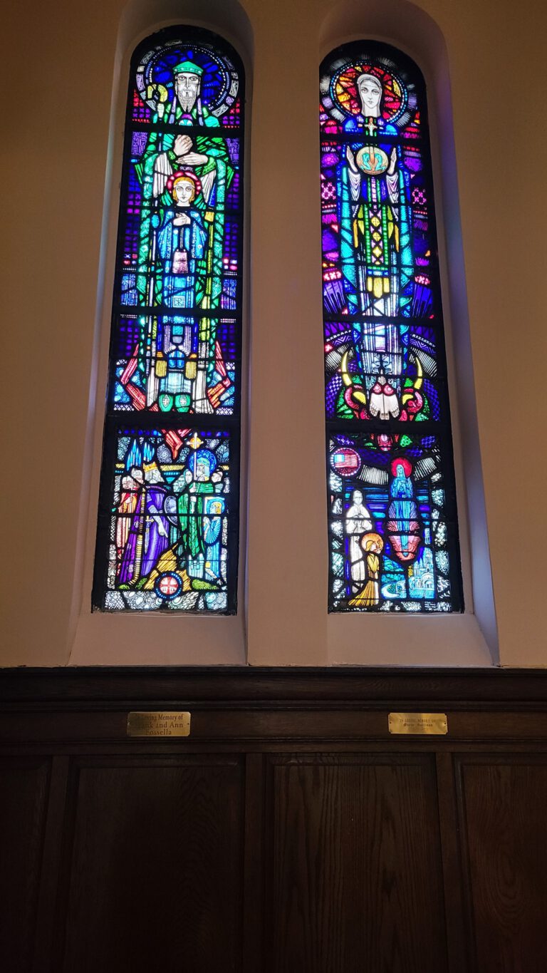 Two tall arched stained glass windows above dark wood paneling in a house of worship