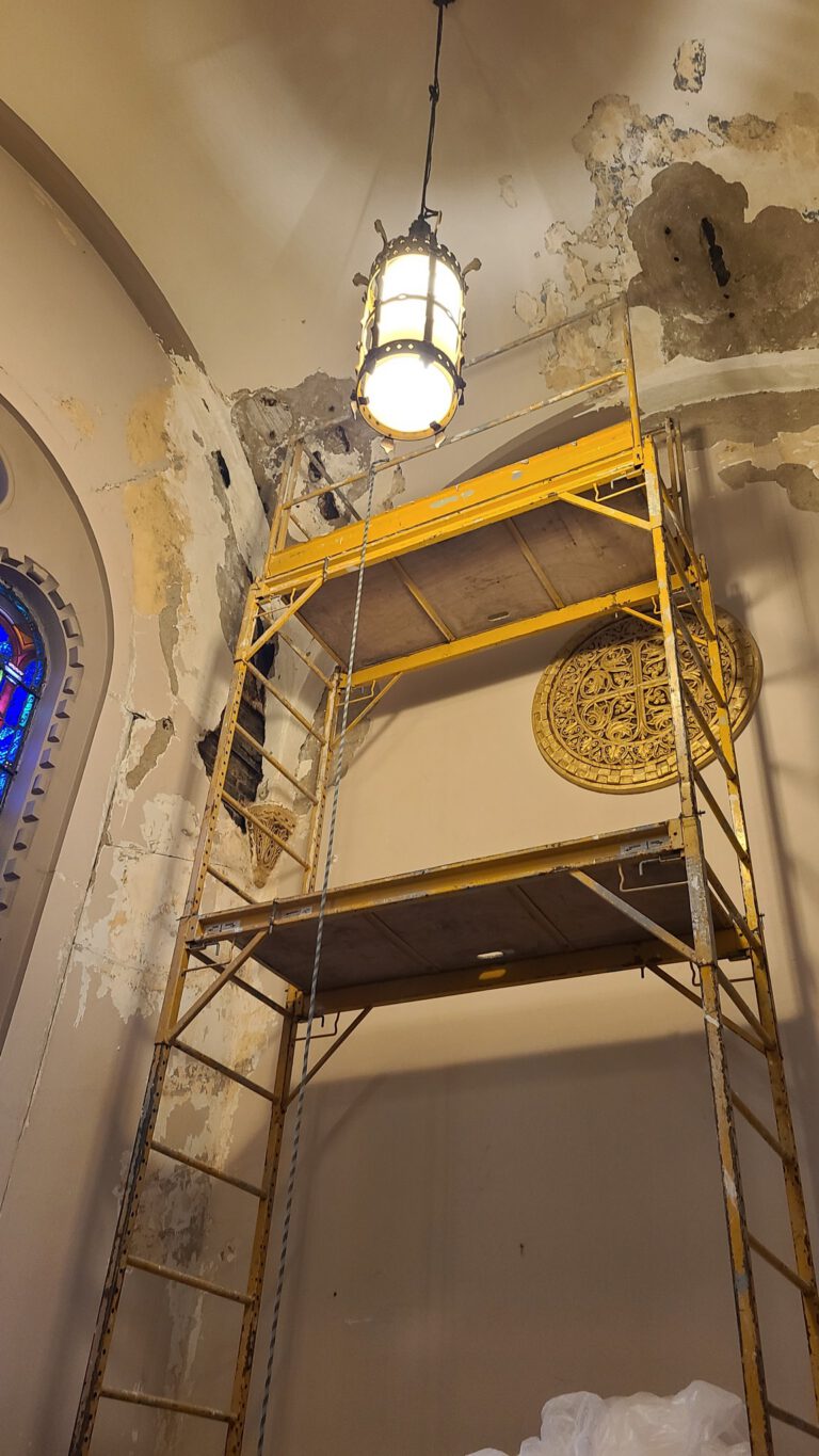 Yellow scaffold tower positioned beside deteriorating plaster wall inside a house of worship